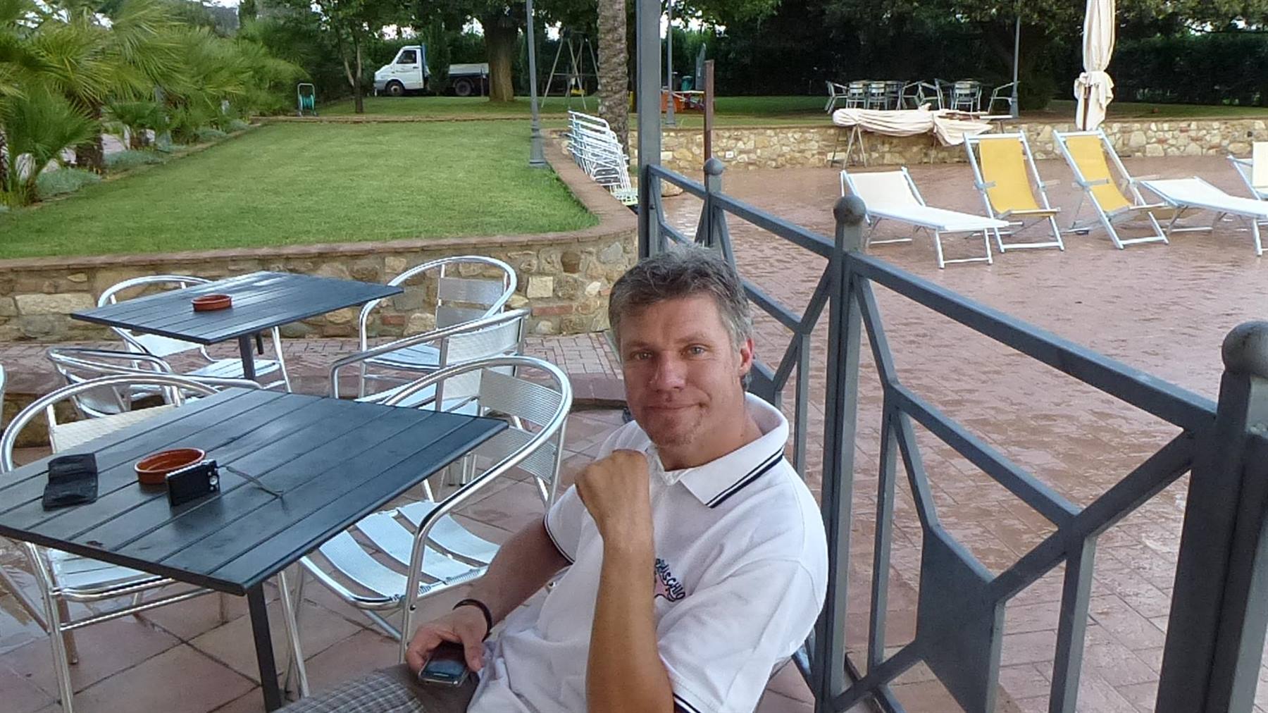 Uomo sorridente seduto in terrazza con tavoli in metallo e sdraio vicino alla piscina