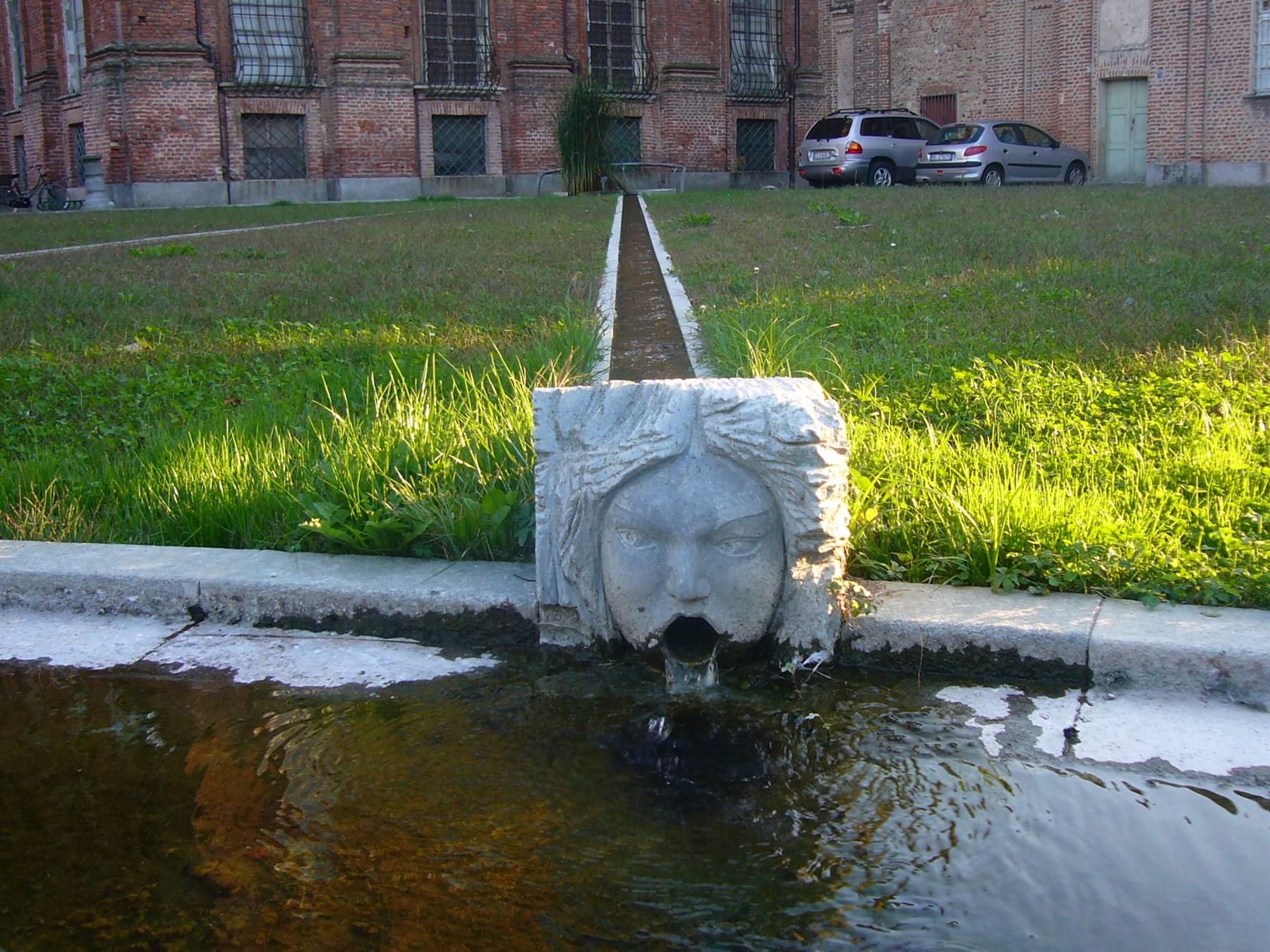 Fontana di pietra con volto scolpito versa acqua, prato e mattoni a Villa Visconti