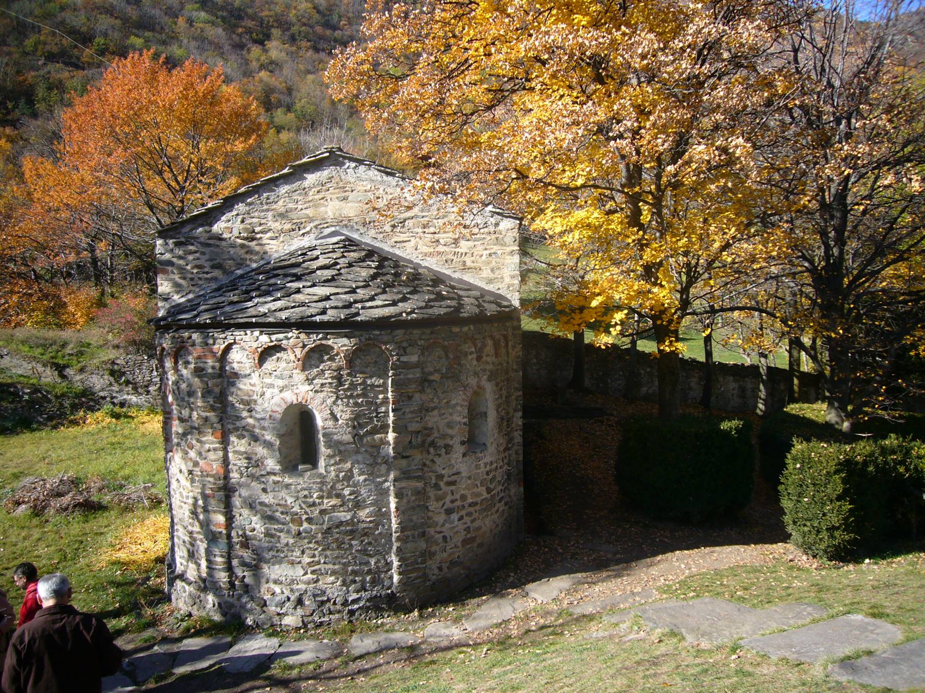 Chiesetta romanica in pietra con abside, tra alberi autunnali, due visitatori