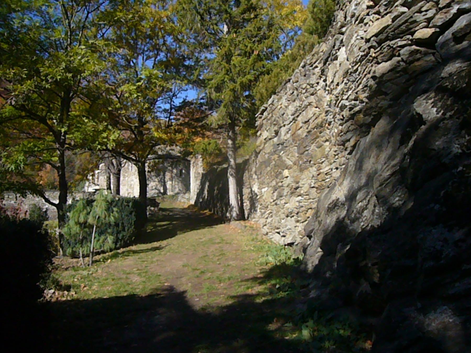 Sentiero erboso tra mura di pietra a secco e alberi autunnali, vecchi edifici in fondo