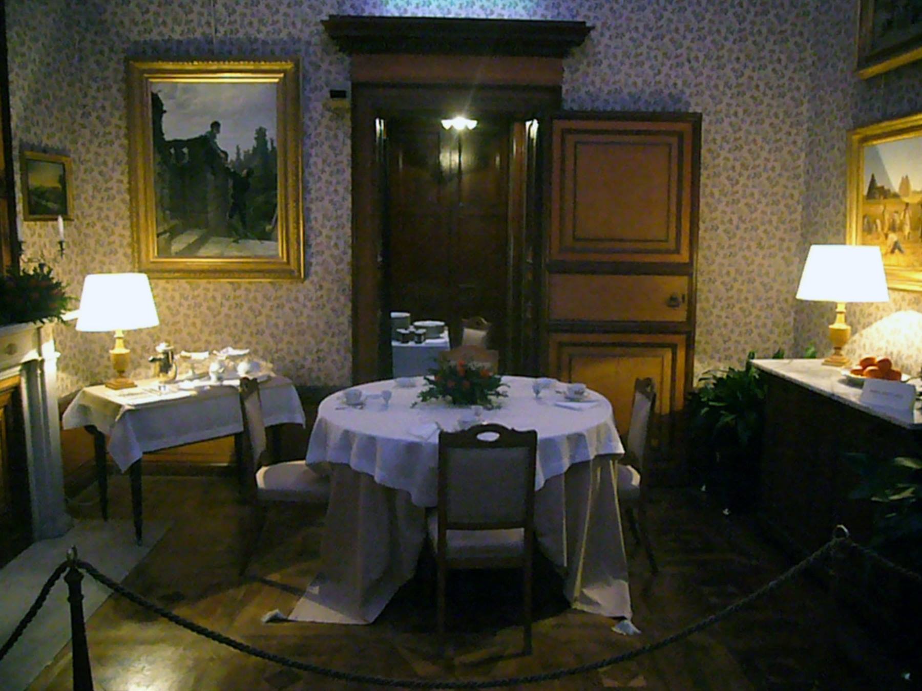 Sala da pranzo con tavolo apparecchiato, lampade accese e quadri dorati alle pareti