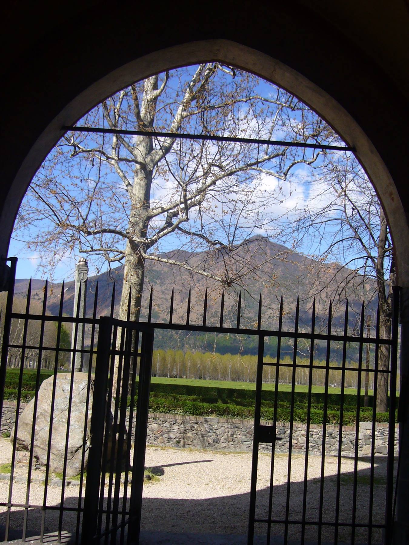 Cancello in ferro sotto un arco di pietra, alberi e montagna sullo sfondo