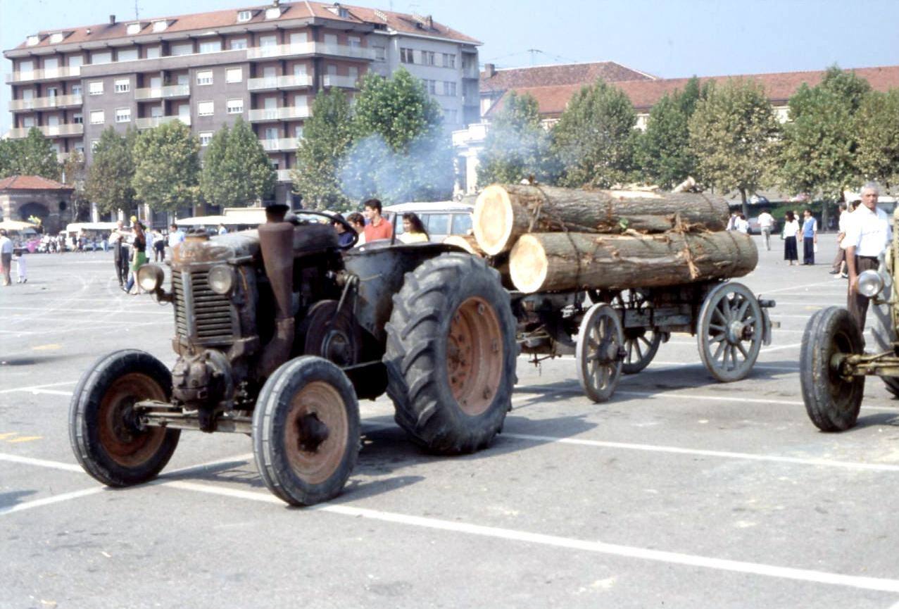 Vecchio trattore d'epoca rimorchia tronchi su un piazzale urbano affollato