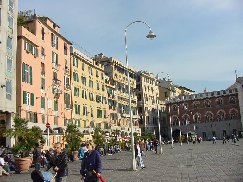 Persone camminano in Piazza de Ferrari a Genova tra palazzi colorati e lampioni curvi