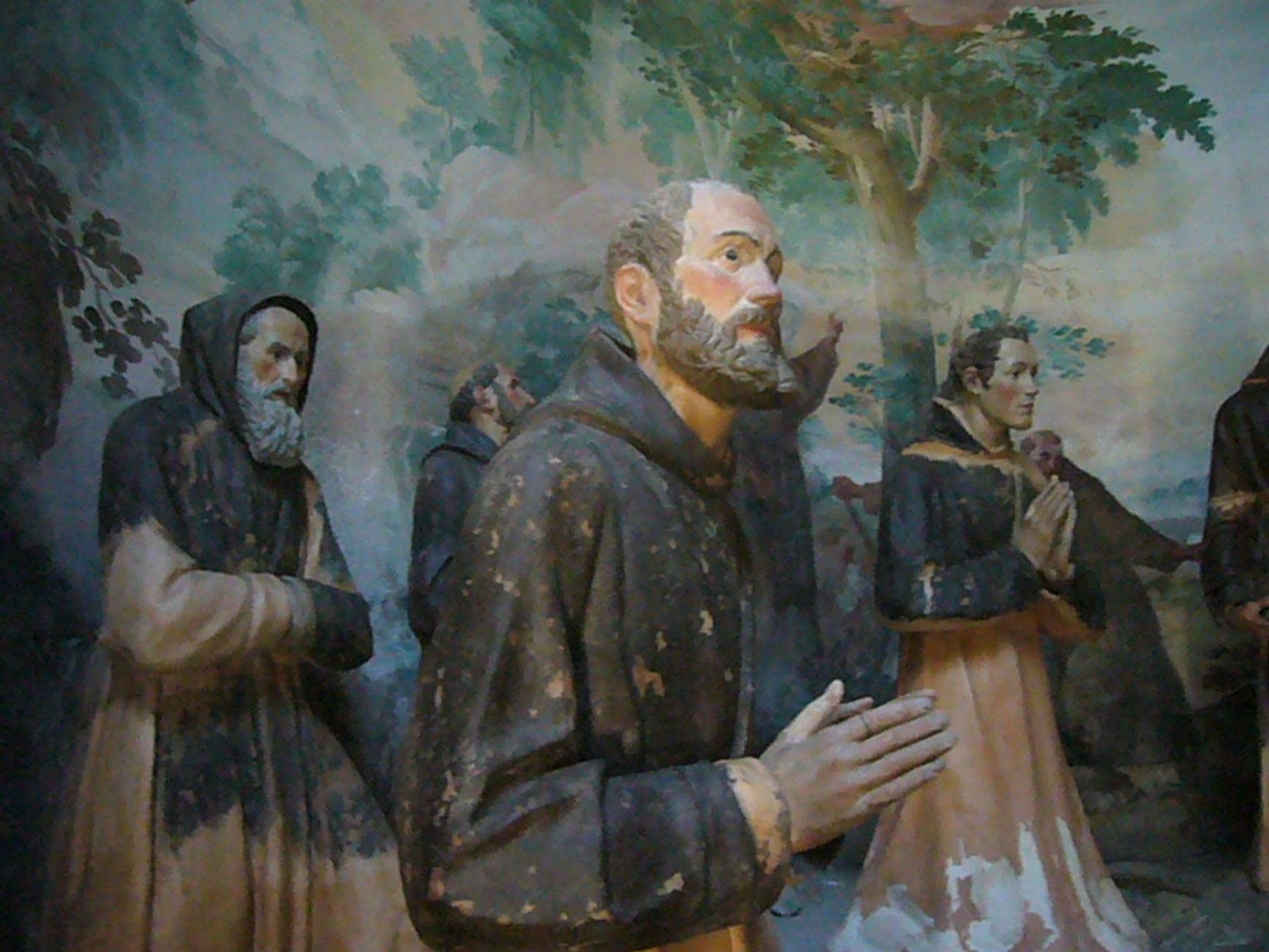 Statue di frati in preghiera, abiti marroni consumati, davanti ad affresco con alberi