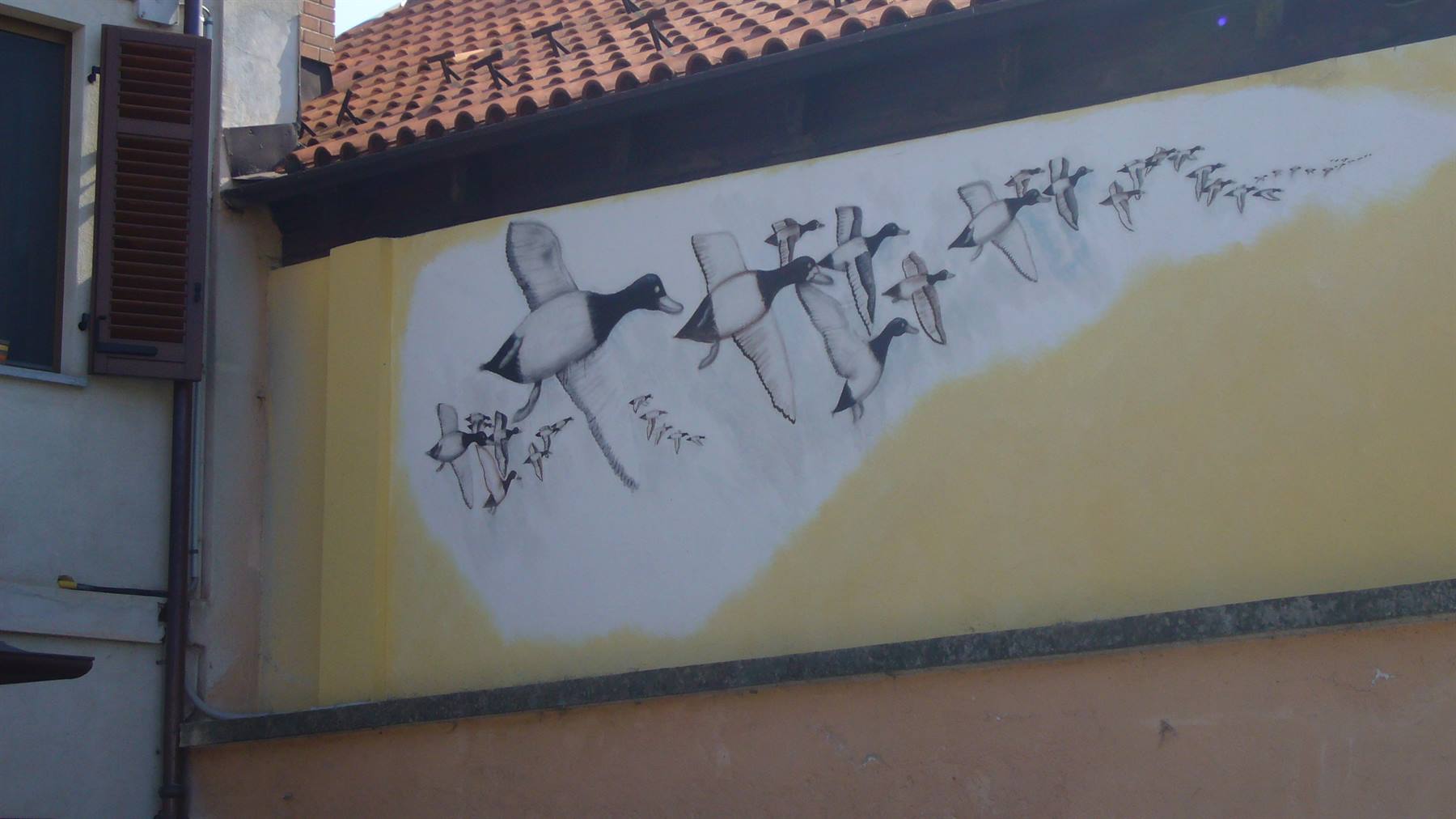 Murale di anatre in volo dipinte su muro giallo sotto tetto in coppi