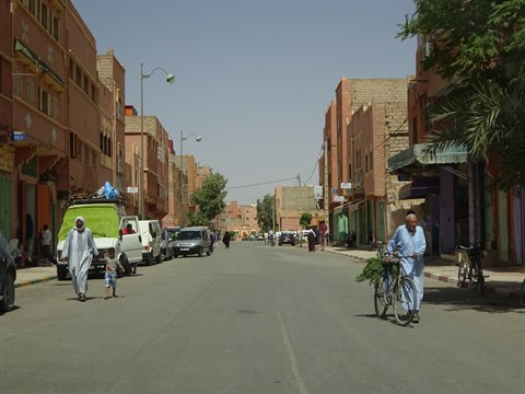 Uomo in djellaba azzurra spinge bicicletta lungo strada ombreggiata di Ouarzazate