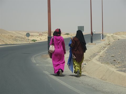 Donne in djellaba camminano lungo l'asfalto tra sabbia e lampioni a Merzouga