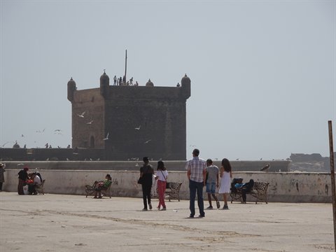 Turisti camminano verso la Sqala du Port a Essaouira, gabbiani nel cielo nebbioso