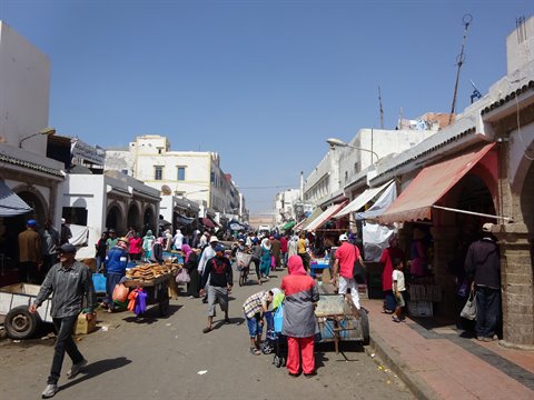 Mercato all'aperto tra edifici bianchi e arcate, gente che passeggia sotto tende colorate, Essaouira