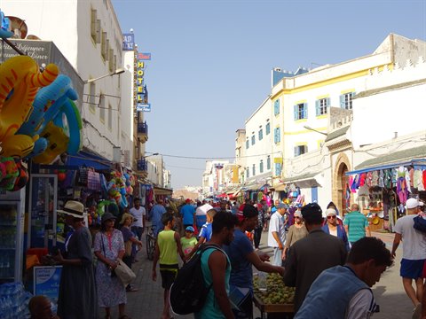 Via affollata di Essaouira tra bancarelle di souvenir e case bianche con persiane azzurre