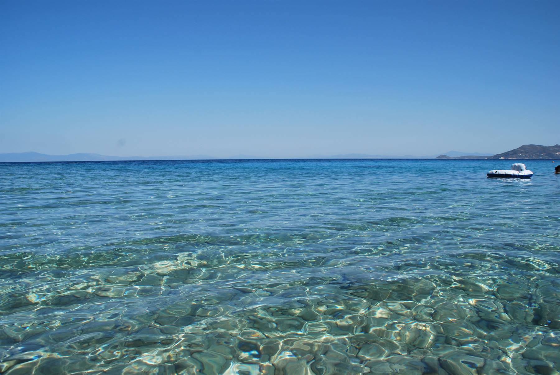 Acqua turchese trasparente sopra sassi visibili, pedalò bianco galleggia, cielo sereno