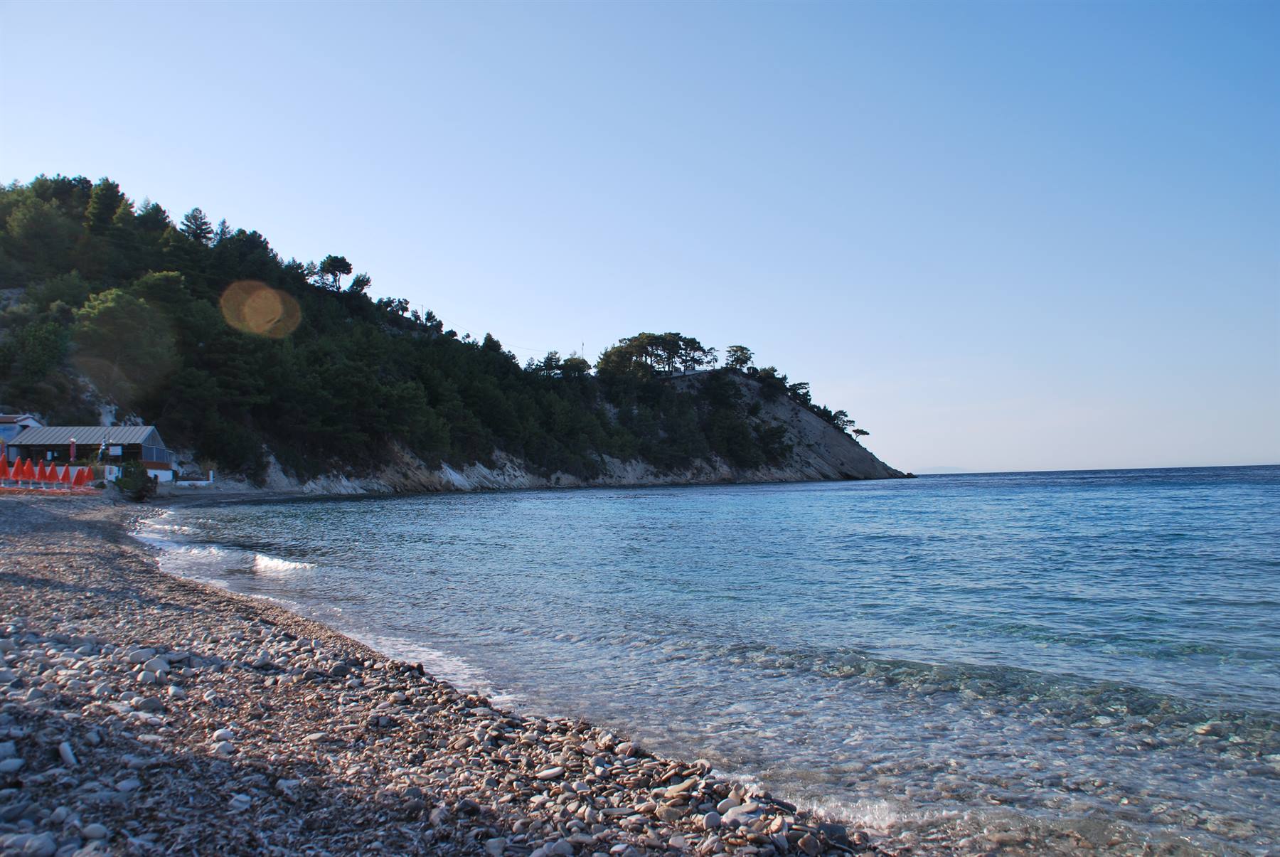 Spiaggia di ciottoli con ombrelloni rossi e mare trasparente sotto collina verde