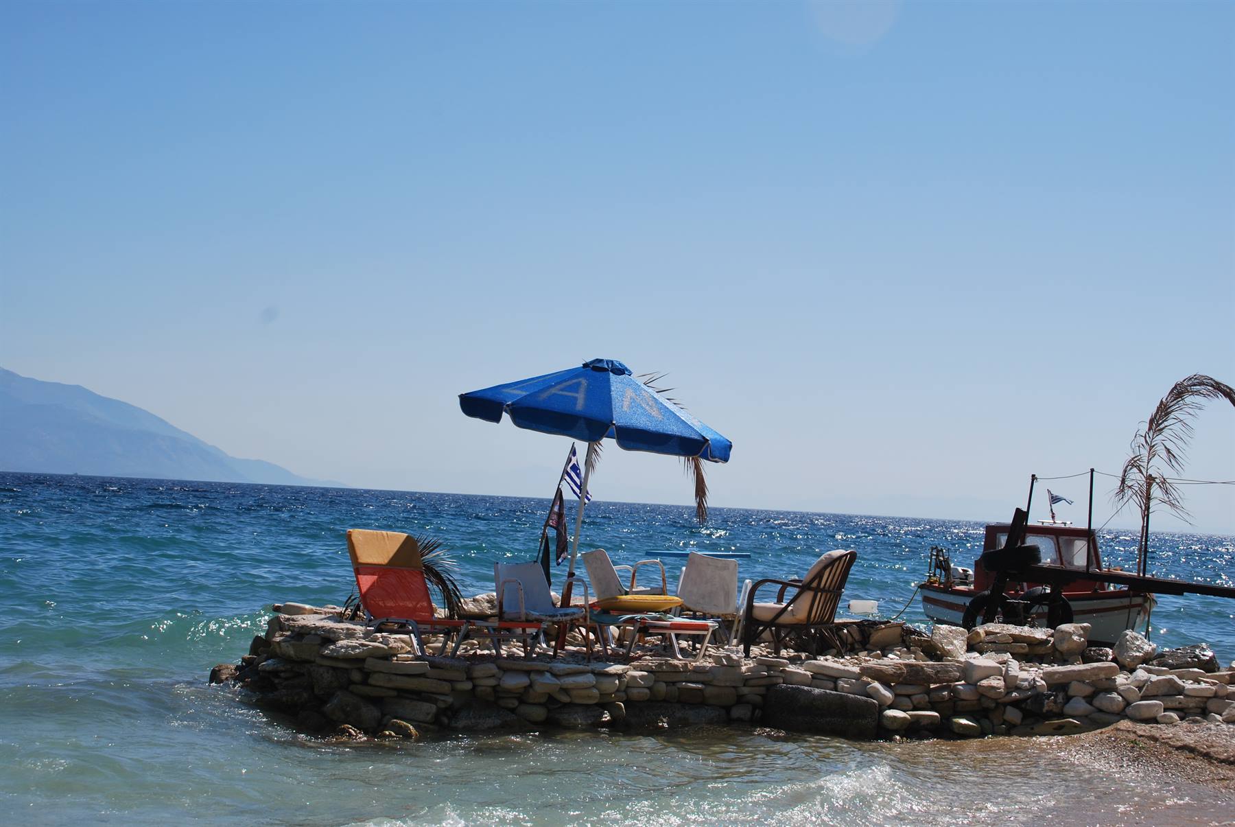 Ombrellone e sedie su molo di pietra, barca greca accosto, mare azzurro