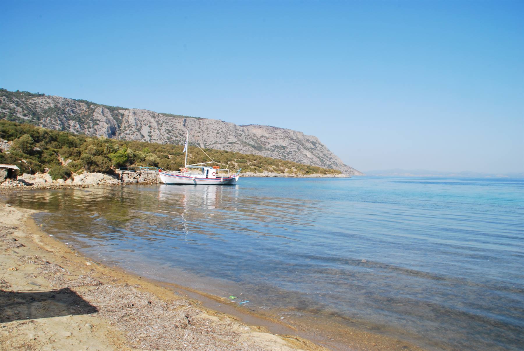 Barca da pesca bianca ormeggiata in baia trasparente, spiaggia sabbiosa, monti aridi sotto cielo azzurro