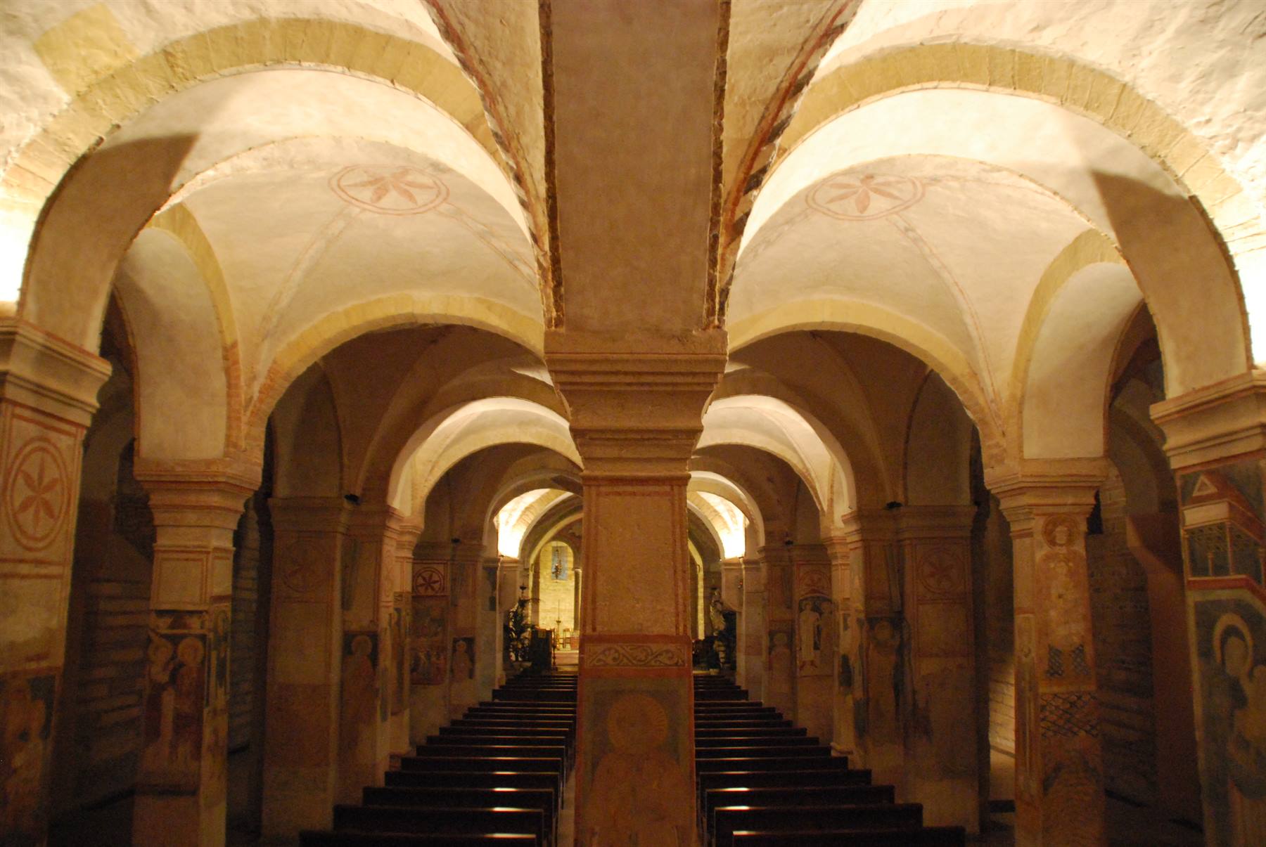 Cripta con volte a crociera, colonne affrescate e banchi di legno in luce calda