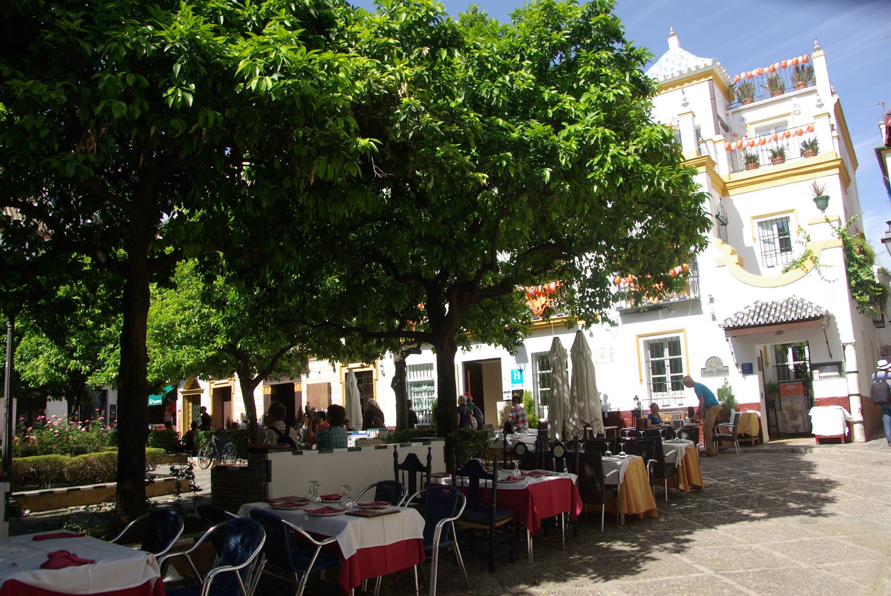 Tavoli all'aperto di un ristorante sotto alberi, edificio bianco e giallo soleggiato