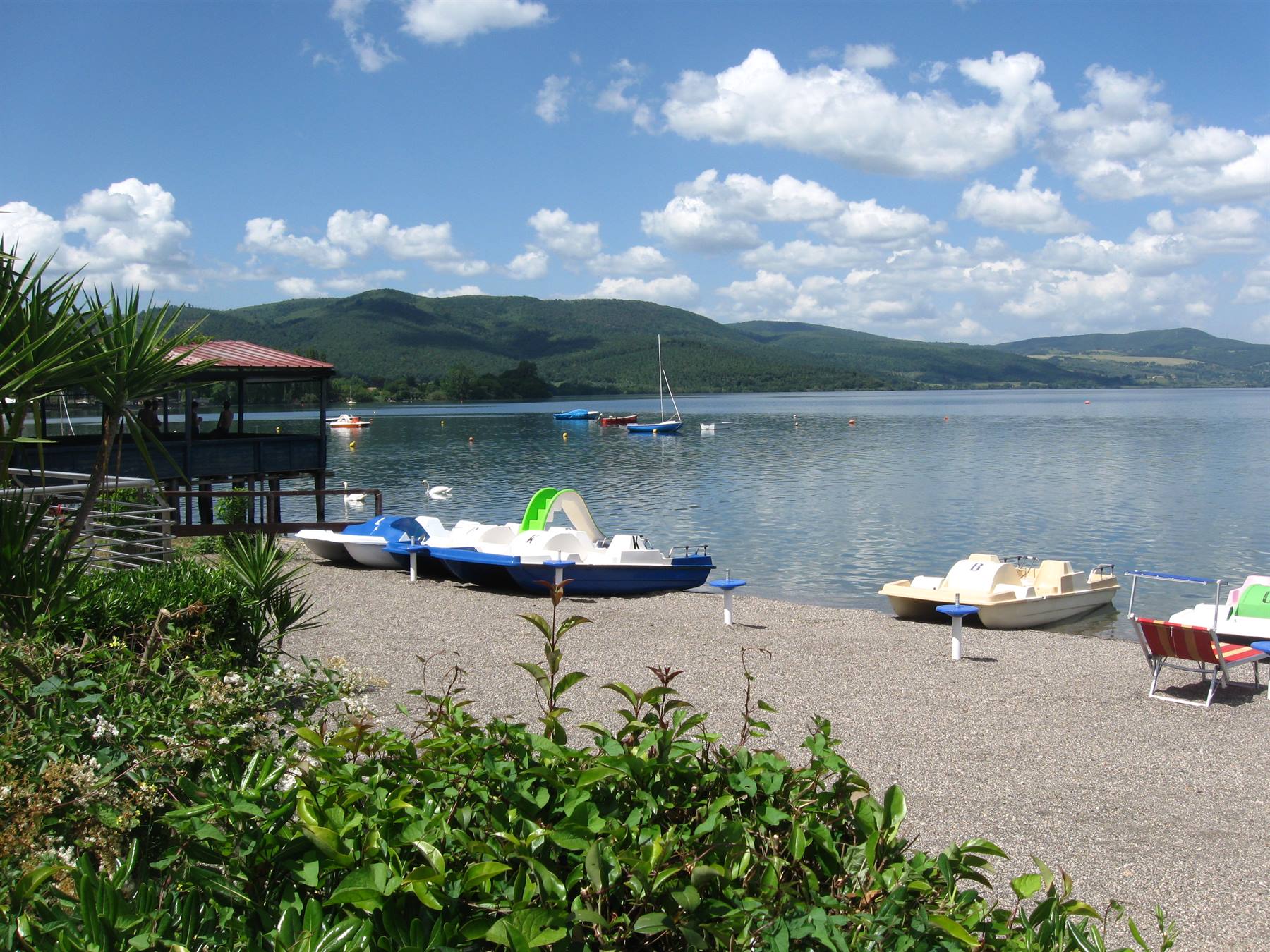 Pedalò colorati sulla spiaggia ghiaiosa del lago, colline e nuvole sullo sfondo