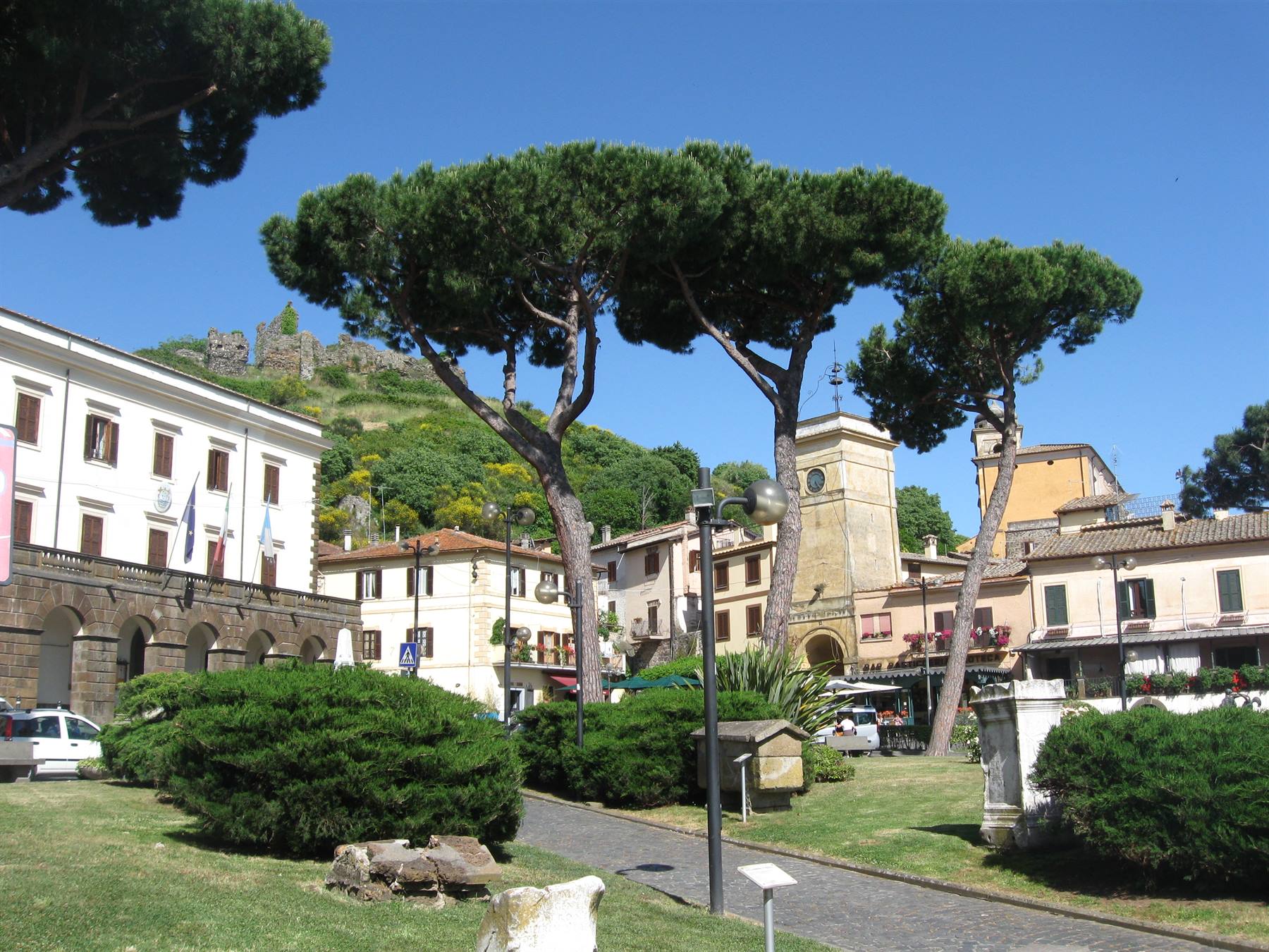Pini alti su piazza con case colorate, torre con orologio e castello sul colle