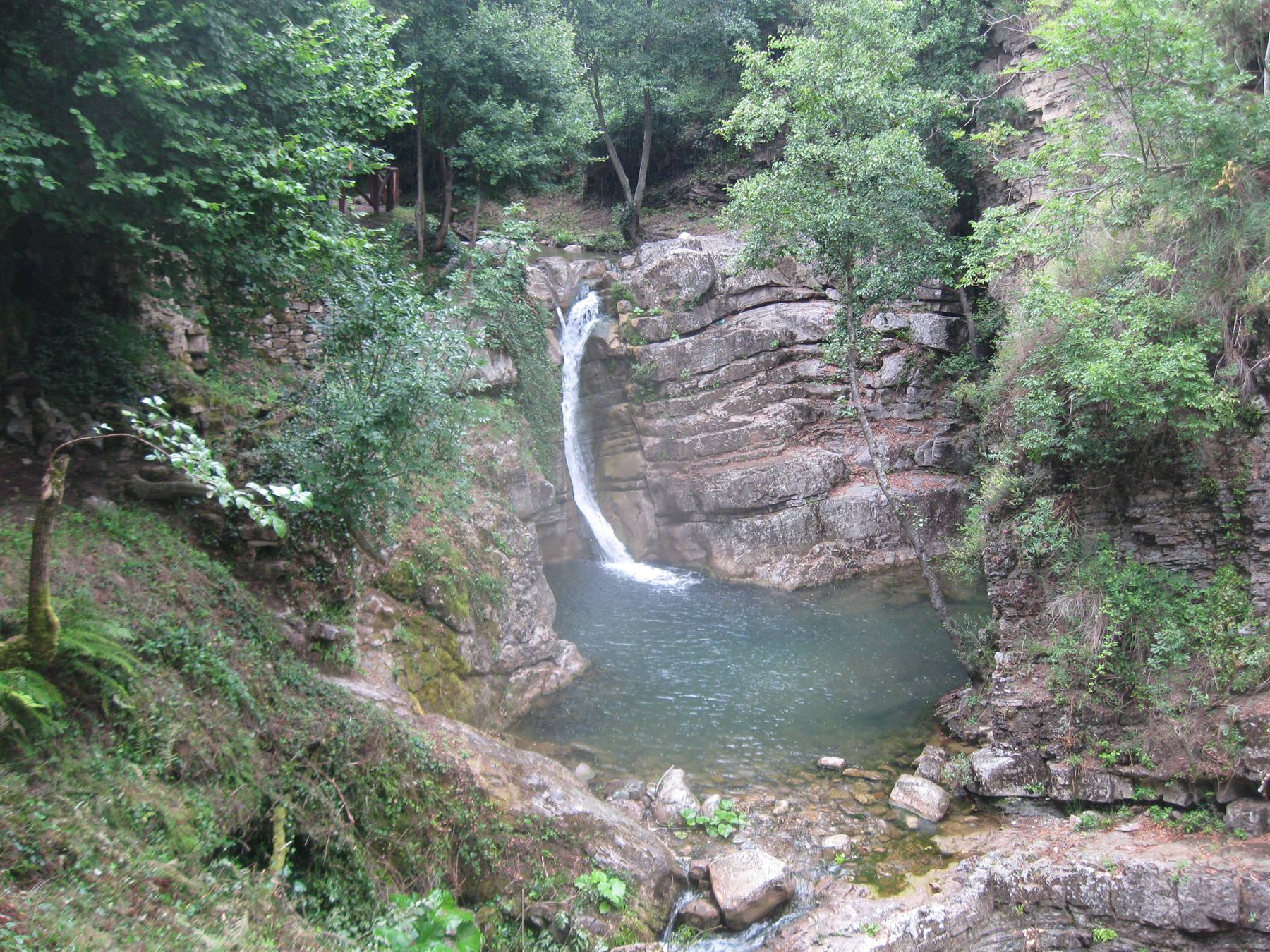 Cascata che scende su rocce stratificate in un laghetto verde circondato da alberi