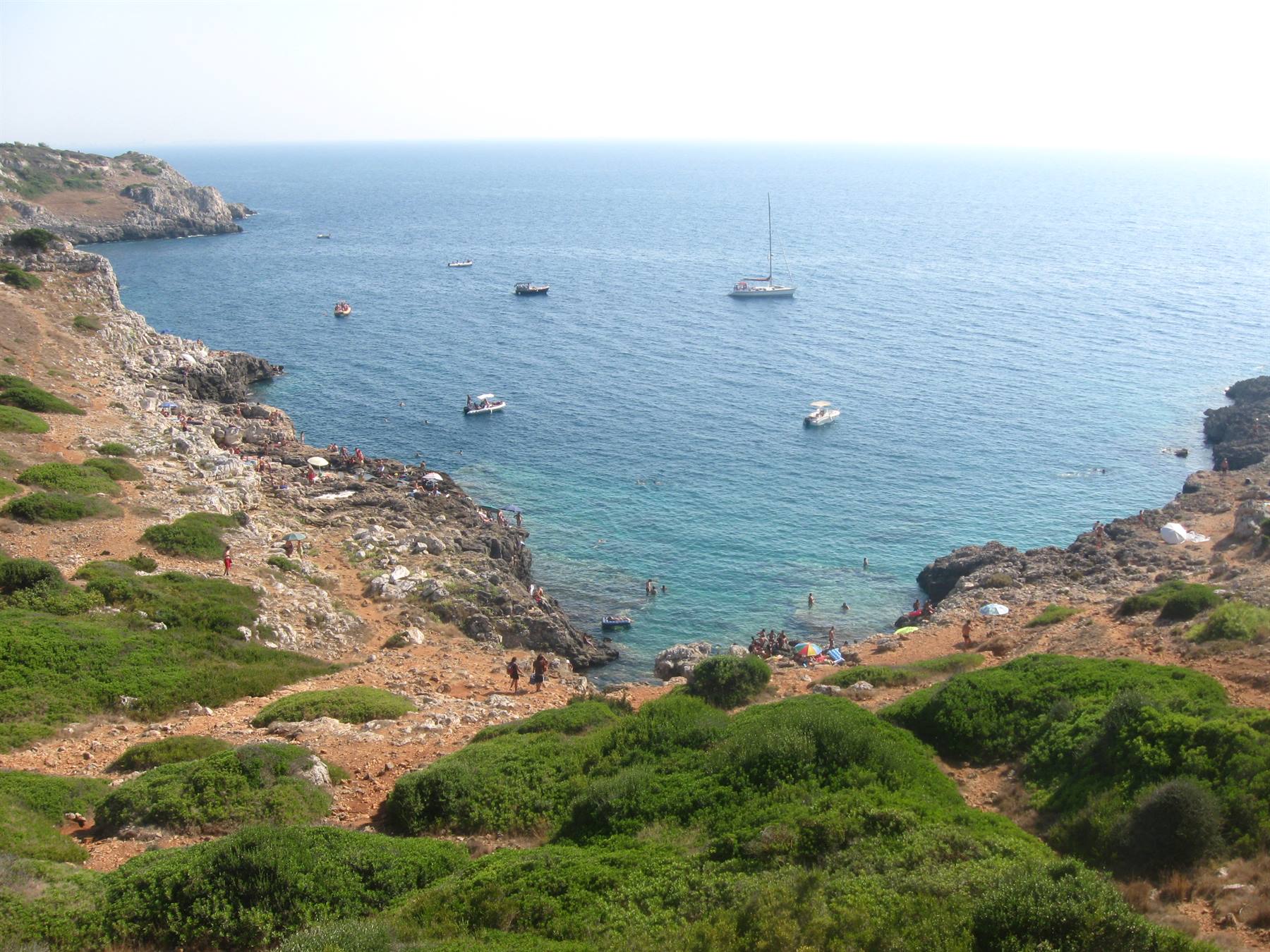 Barche in una caletta rocciosa con scogliera, macchia mediterranea e bagnanti