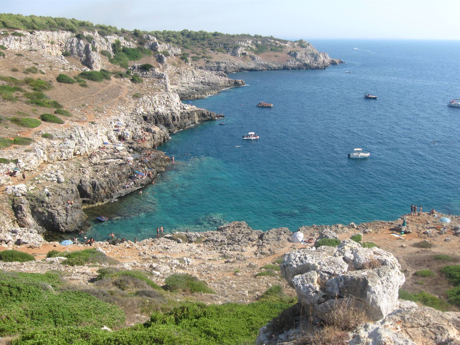 Cala rocciosa con acque turchesi e barche, scogliera calcarea salentina con bagnanti