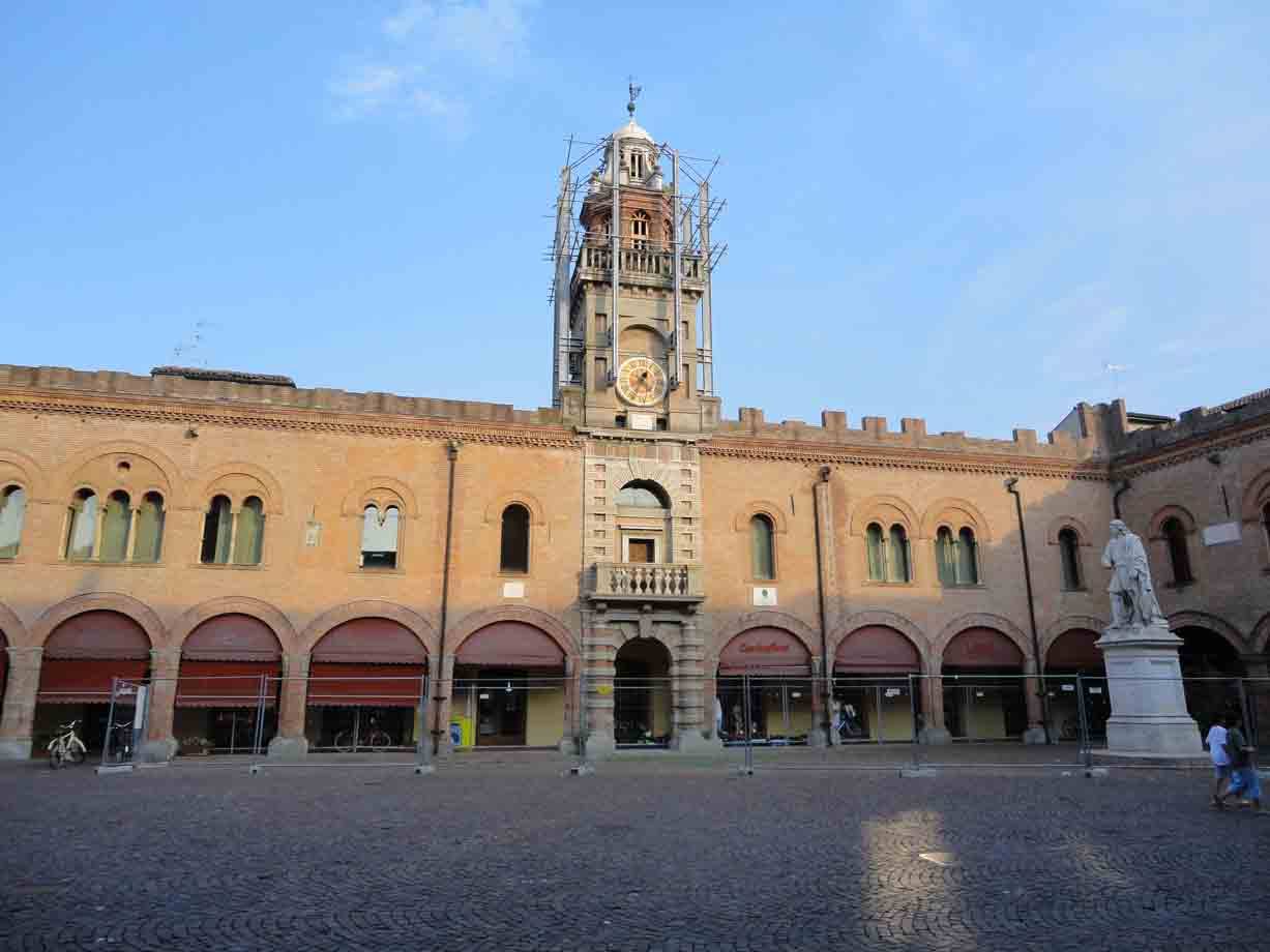 Piazza con palazzo in mattoni, torre dell'orologio in restauro e statua