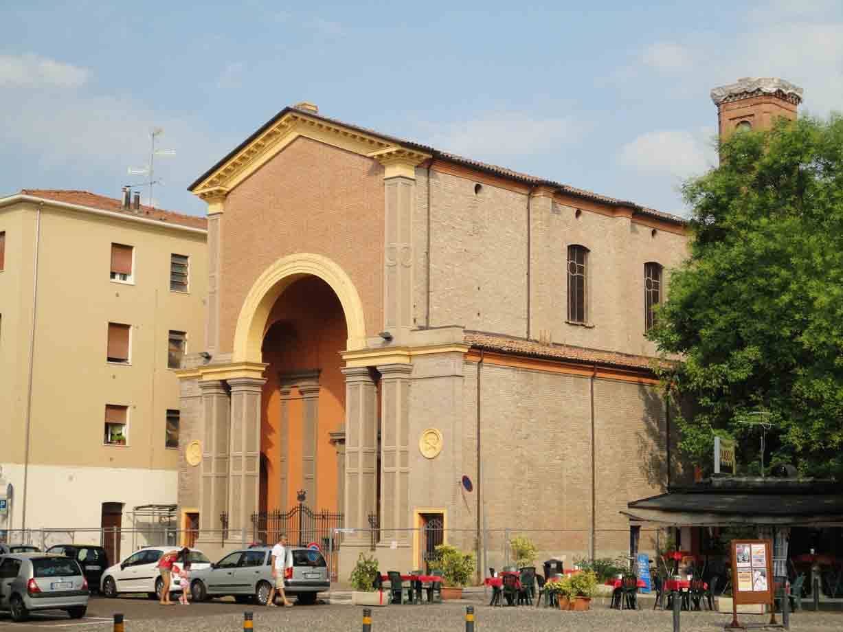 Facciata in mattoni con grande arco, chiesa affacciata su piazza con auto e caffè