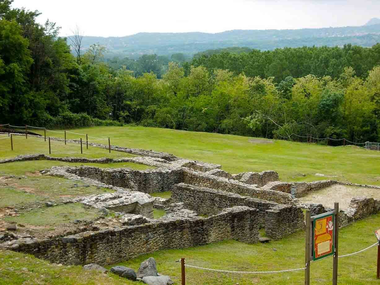 Rovine archeologiche in pietra tra prati verdi e bosco, colline sullo sfondo
