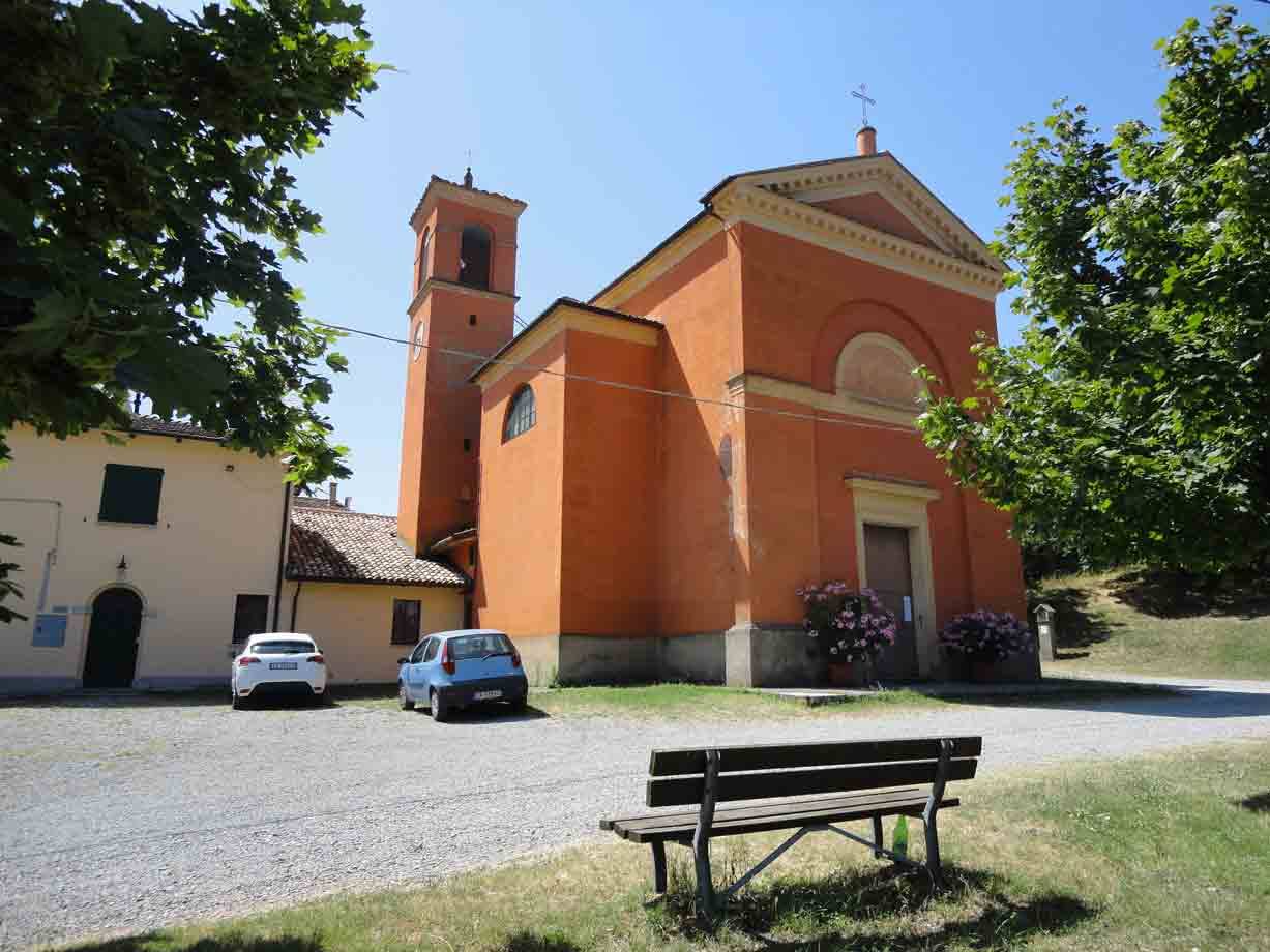 Chiesa arancione con campanile, panchina in legno e auto parcheggiate sotto il sole