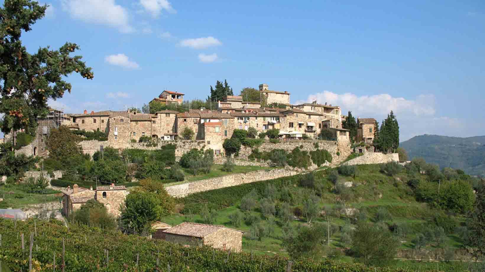 Borgo di case in pietra su collina, vigneti e ulivi sotto, cielo azzurro