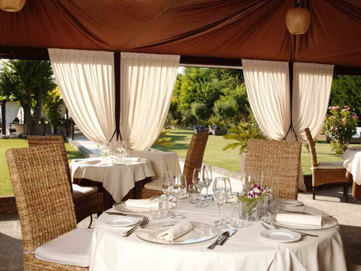 Tavolo apparecchiato con bicchieri e sedie in vimini sotto tenda con tendaggi bianchi
