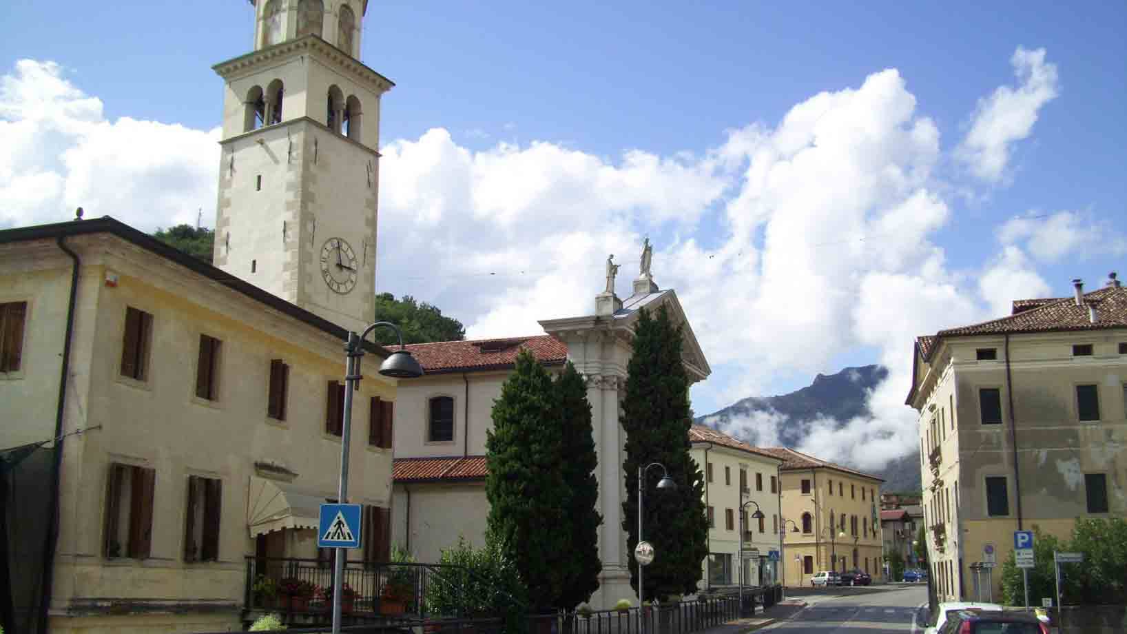 Campanile e chiesa tra case gialle in piazza, monti nuvolosi sullo sfondo