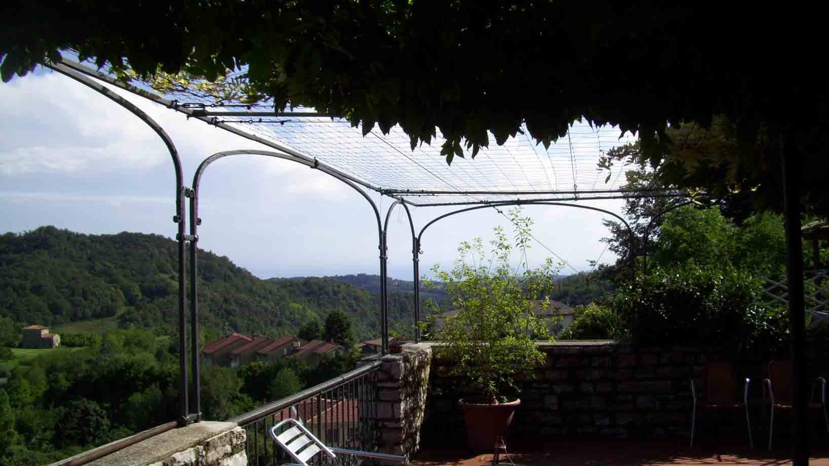 Terrazza con pergolato di ferro e viti su colline verdeggianti, sedie in metallo vuote