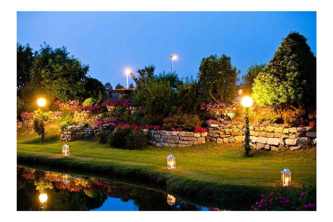 Giardino fiorito con muretti in pietra, lanterne accese e luci riflesse nello stagno