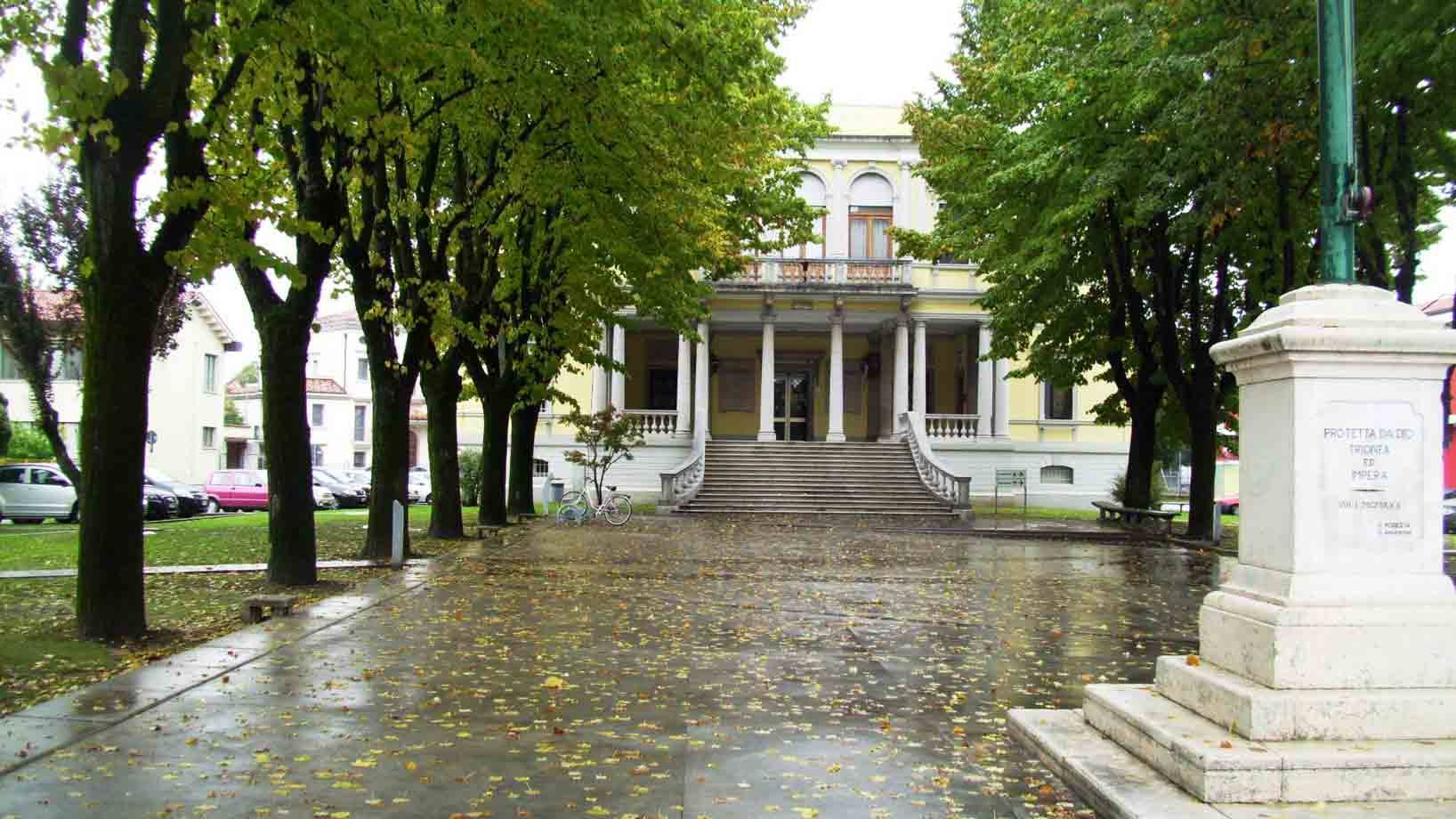 Viale alberato bagnato dalla pioggia verso una villa neoclassica con scalinata, monumento in marmo a destra