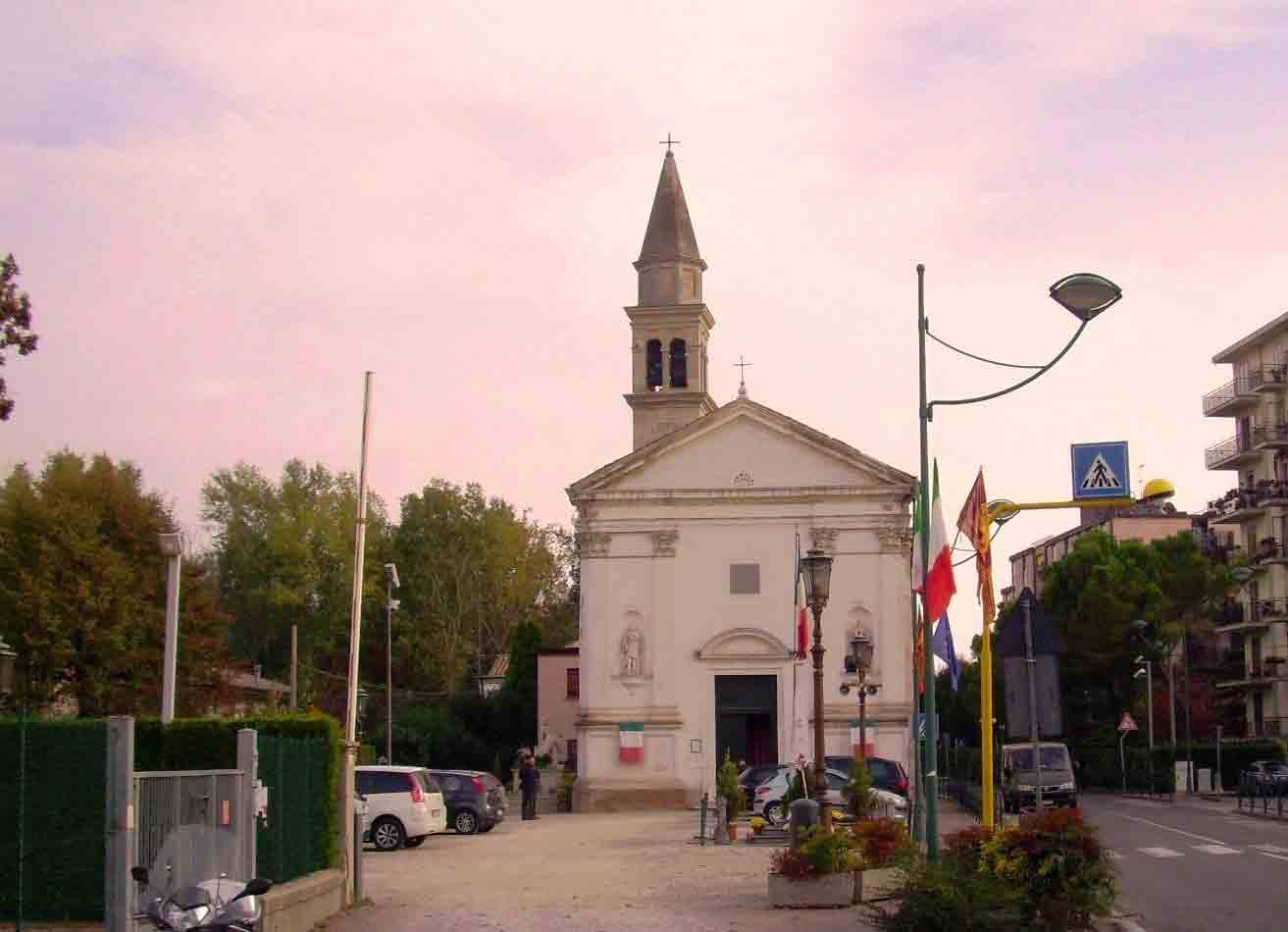 Chiesa bianca con campanile tra auto e bandiere, cielo rosa al mattino