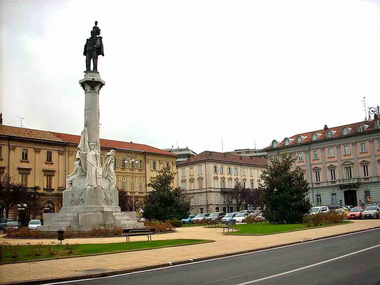 Monumento con statua in bronzo su colonna, piazza con edifici ottocenteschi e auto parcheggiate