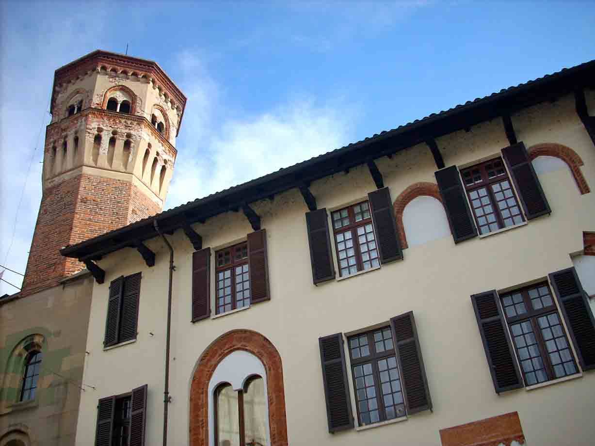 Torre mattone medievale accanto a palazzo crema con persiane, sotto cielo azzurro