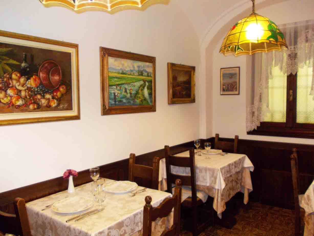 Sala di trattoria con tavoli apparecchiati, quadri alle pareti e lampada gialla
