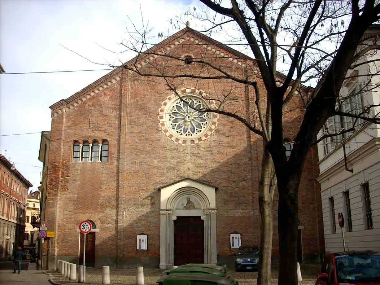 Facciata in mattoni di chiesa con rosone, rami spogli, auto parcheggiate e cielo nuvoloso