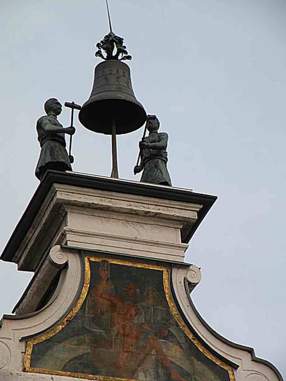 Due statue in bronzo martellano una campana sul timpano di un palazzo affrescato