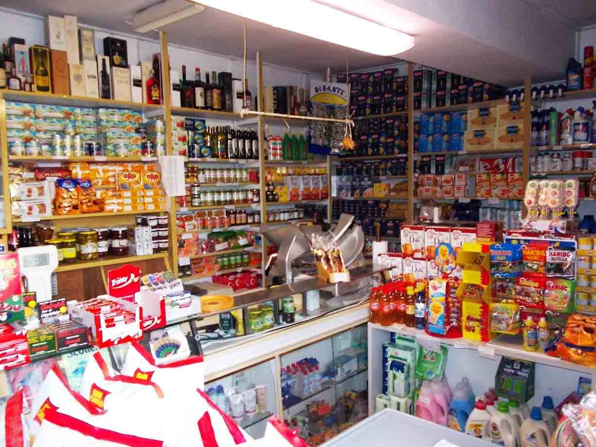 Interno di un piccolo alimentari affollato di scaffali con vini, conserve e detergenti