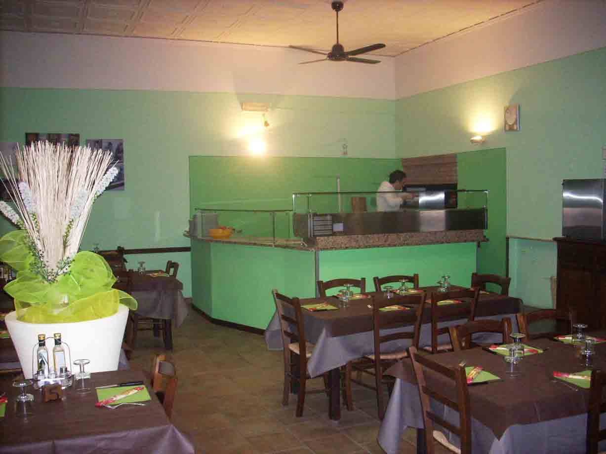 Sala di pizzeria con tavoli apparecchiati, bancone verde e pizzaiolo al lavoro