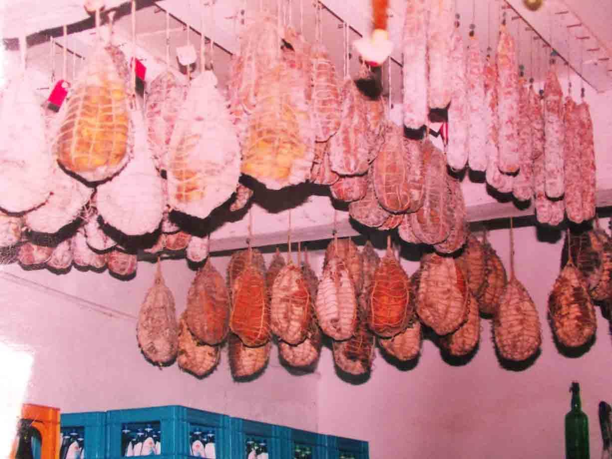 Salumi stagionanti appesi alle corde in una cantina, pareti bianche, luce calda