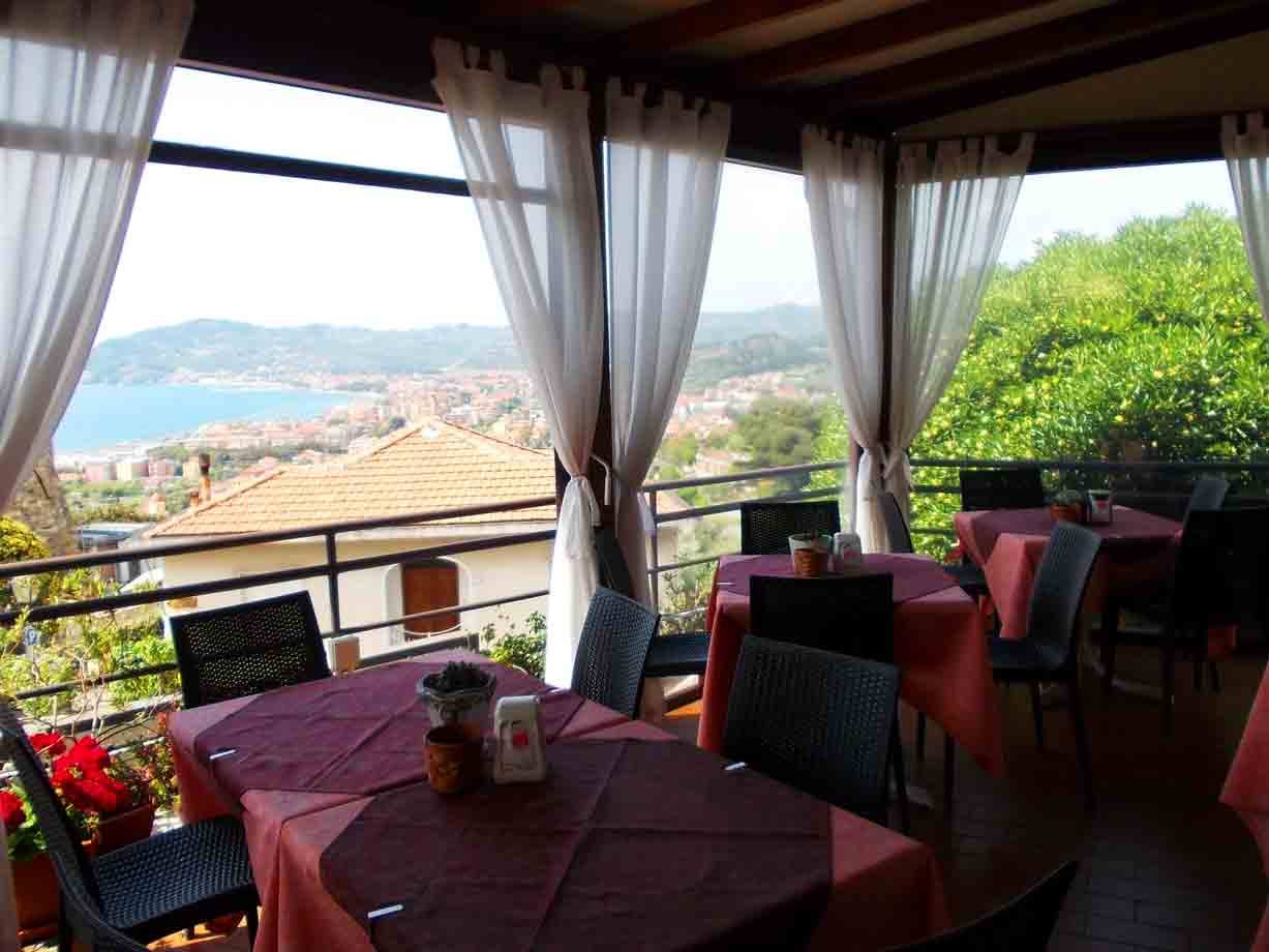 Veranda di ristorante con tavoli apparecchiati e teli bianchi, golfo e tetti affacciati