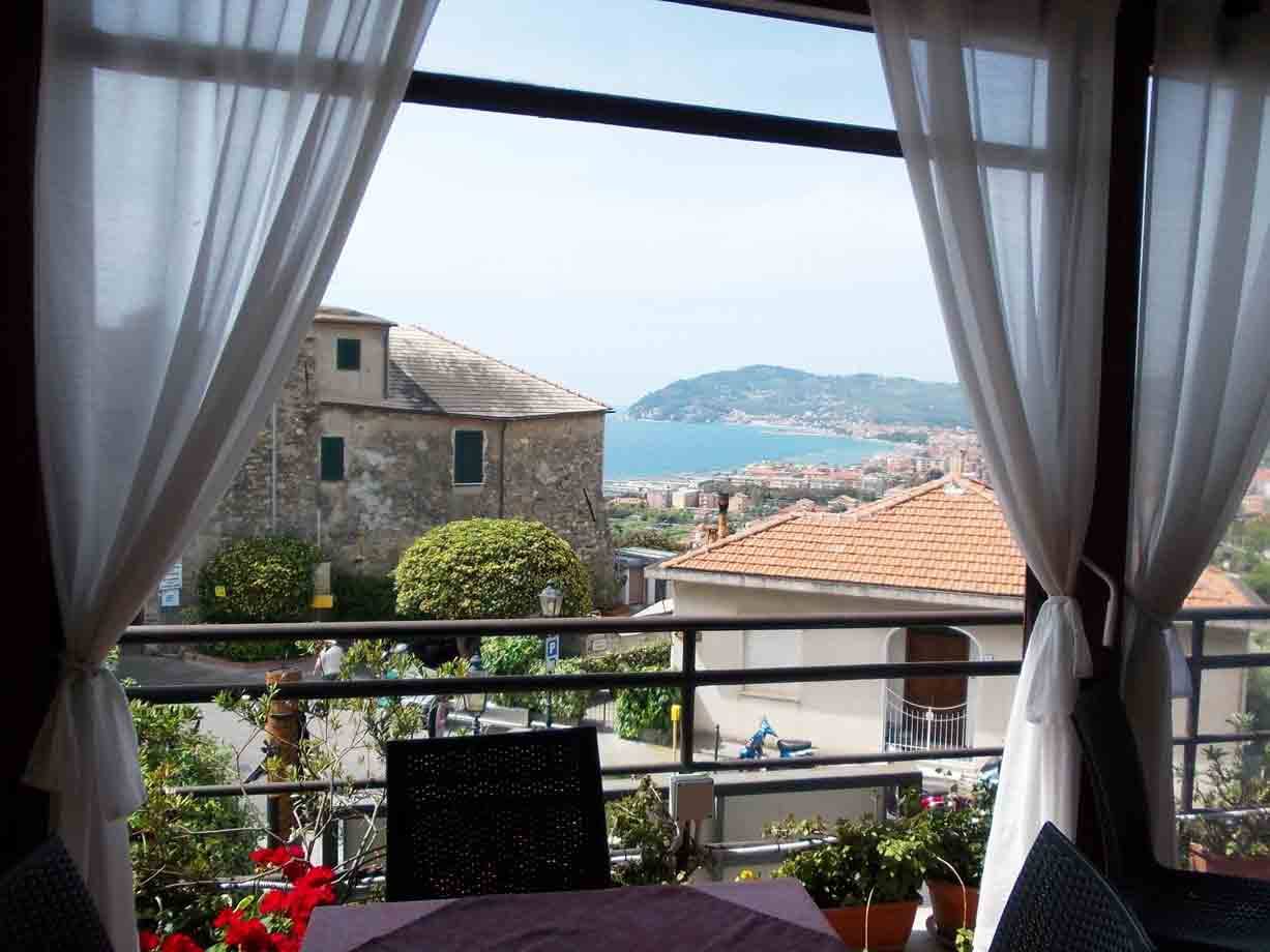 Tavolo in veranda con tende bianche, case in pietra e baia azzurra sullo sfondo