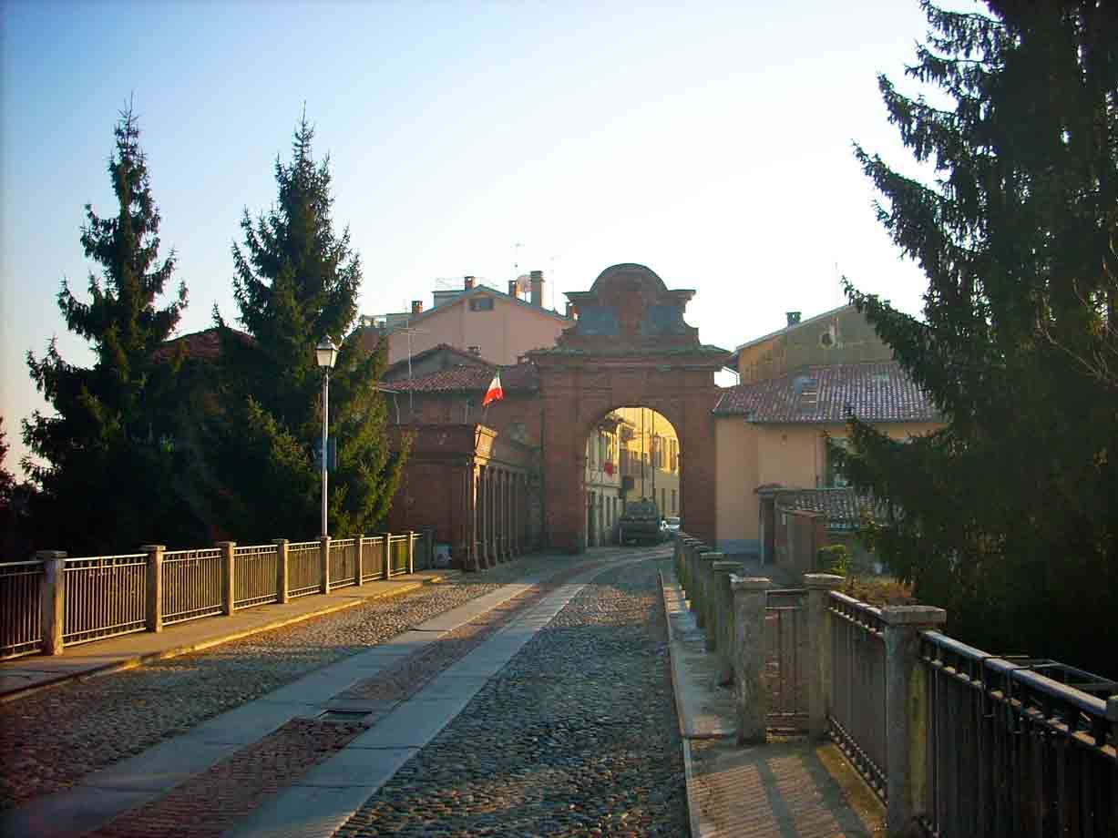 Porta in mattoni alla fine di un ponte lastricato, abeti alti e luce mattutina