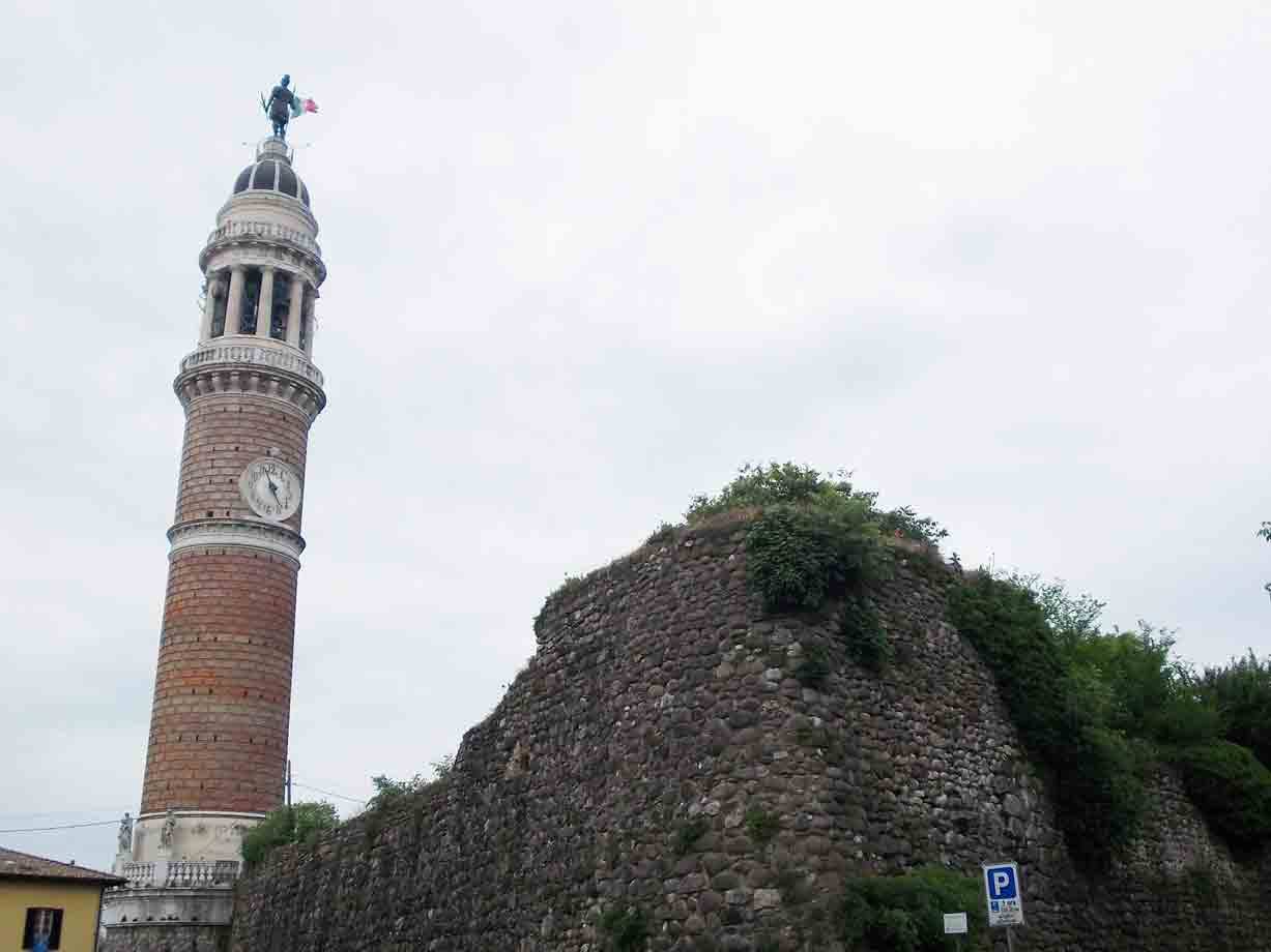 Campanile in cotto con orologio e statua, accanto a ruderi di mura in pietra