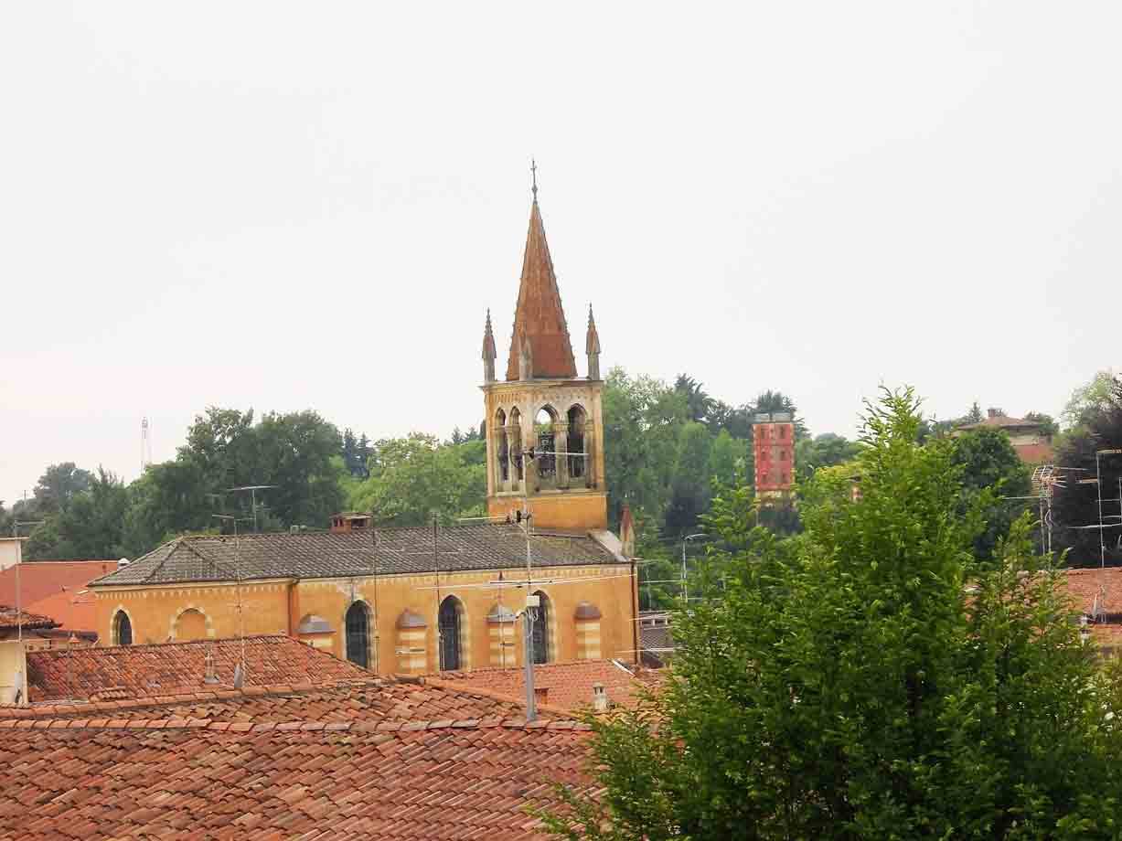 Campanile giallo con guglia tra tetti di coppi e alberi verdi, cielo grigio